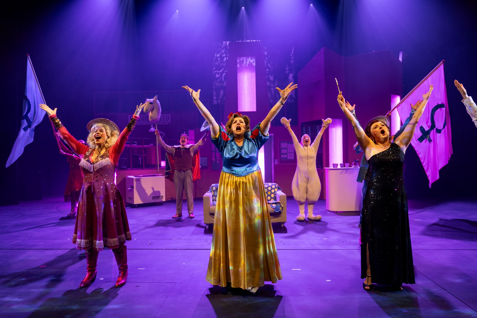 Kuva teatterin musikaaliesityksestä. Kolme naista kuvan etualalla kädet ja katse kohottetuina ylös päin. Yhdellä naisista on iltapuku, yhdellä prinsessa mekko ja yhdellä cowboy-asu. Naisten takana liehuu kaksi lippua, joissa on naaras-symboli, sekö prinssiksi pukeutunut mies ja jänikseksi puketunut mies. 